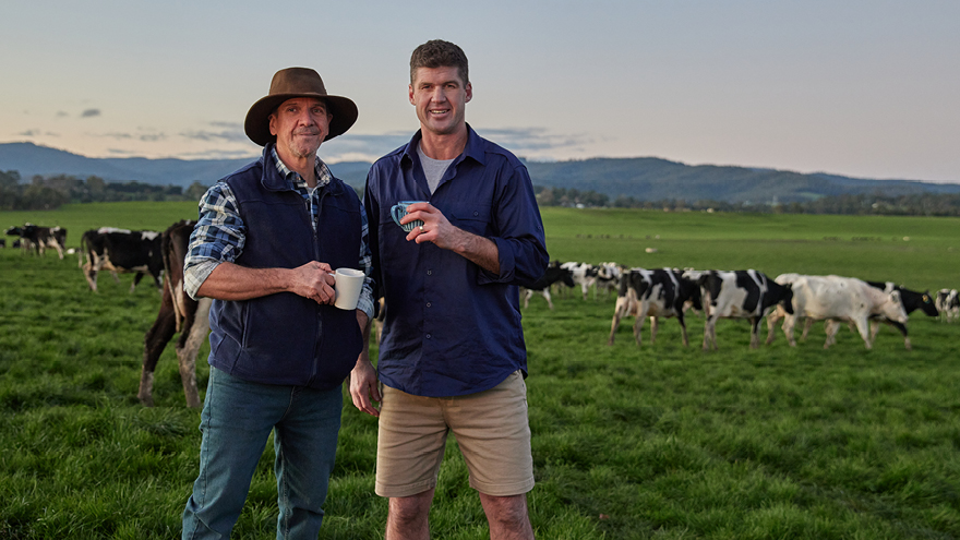 Jonathan Brown and farmer holding glasses of milk. Standing in paddock, cows behind them.