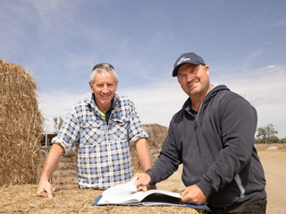 Two male dairy farmers stand beside a hay bale under blue skies. They are smiling, one is holding a report.